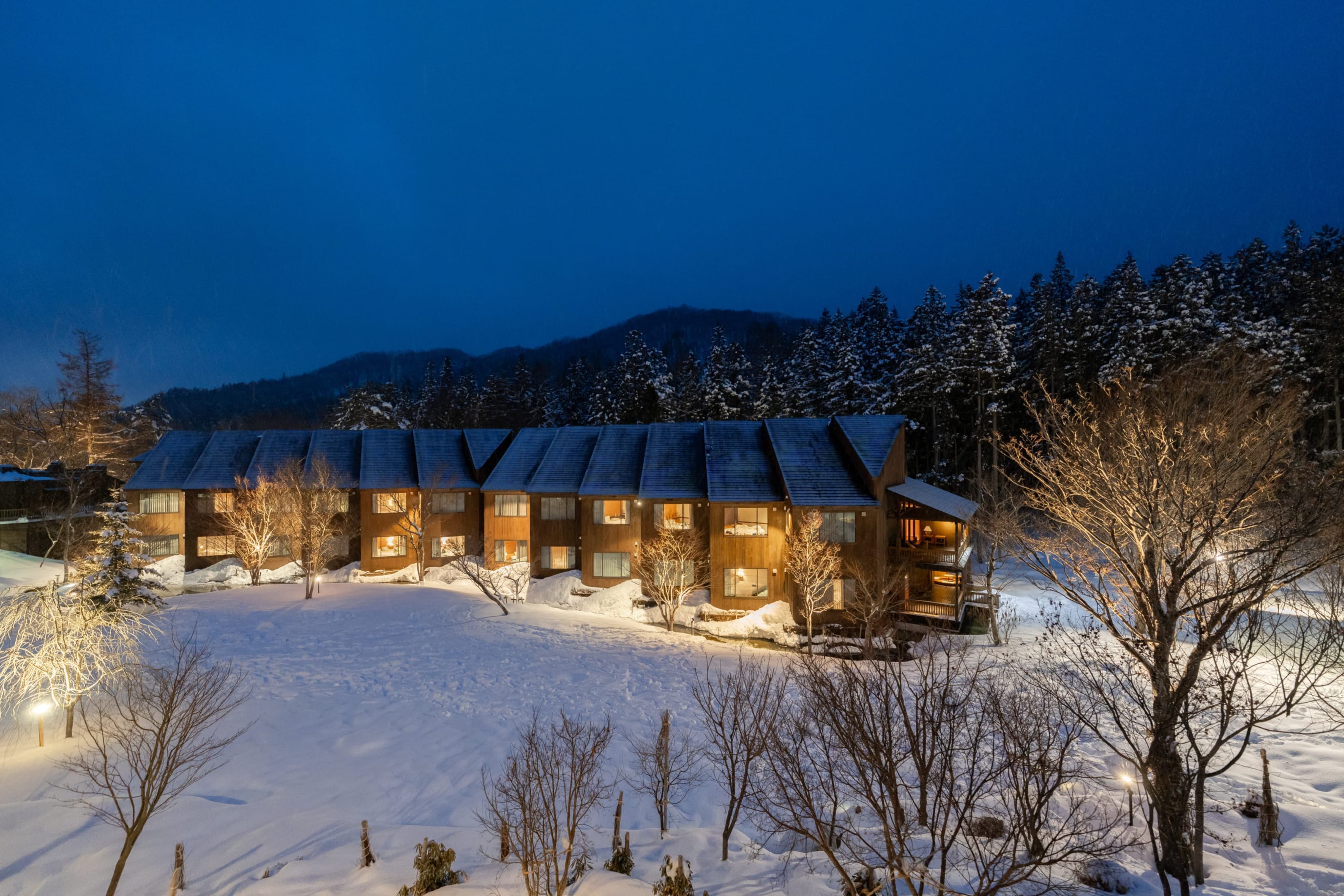 Hotel Sierra Resort Hakuba wood-clad lodge exterior glowing at dusk amid deep snow and forest, Hakuba