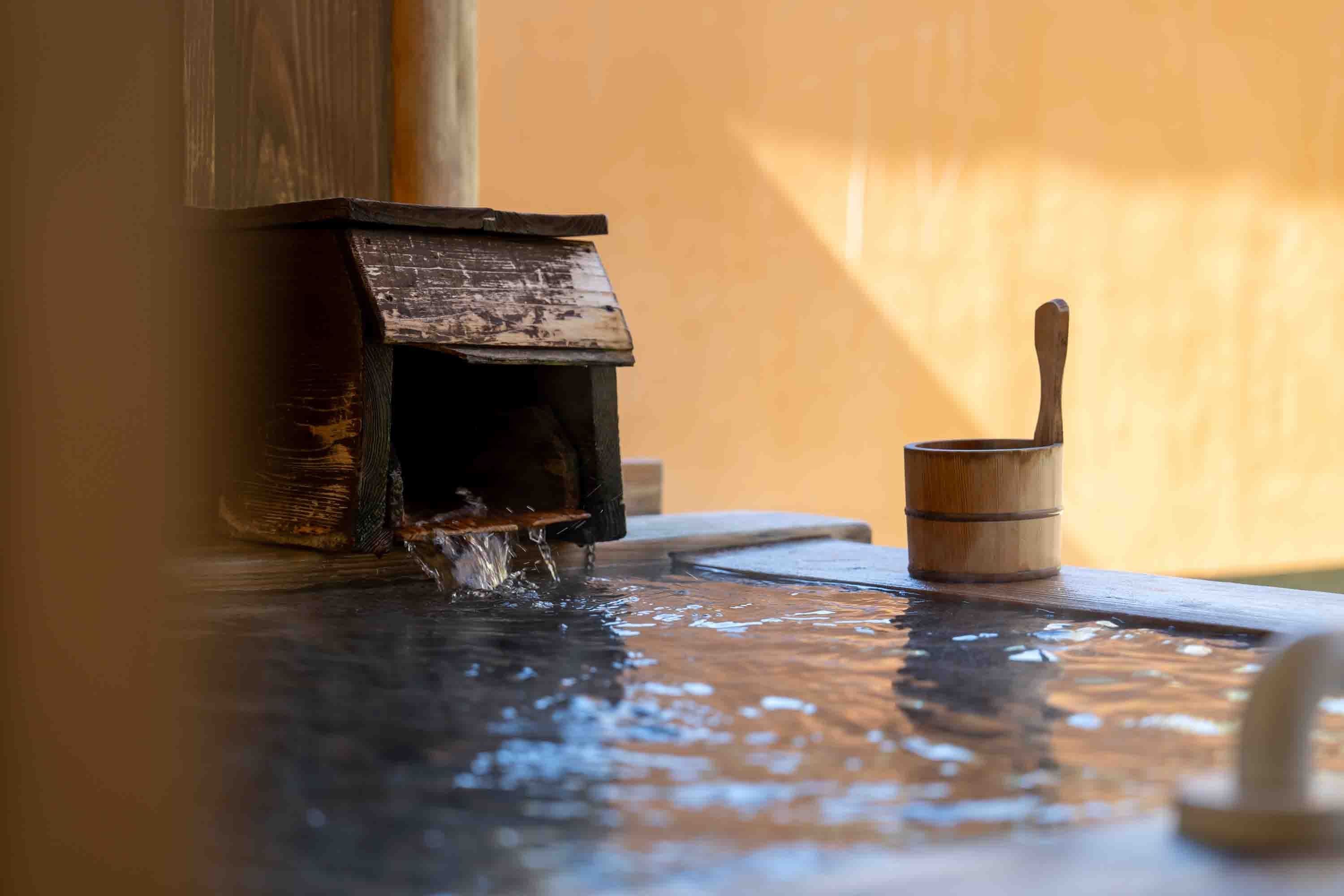Hot-spring water flowing from a wooden spout into a bath beside a cypress ladle at Hotel Sierra Resort Hakuba