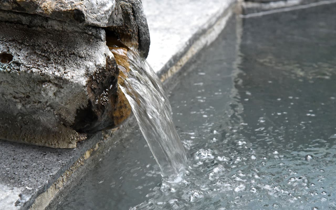 Close-up of hot spring water flowing from a mineral-encrusted rock spout into the bath