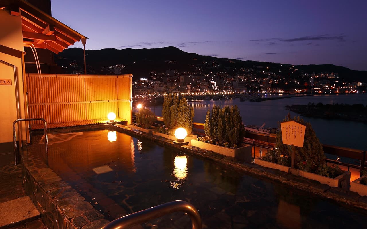Rooftop open-air rotenburo lit by lanterns at dusk overlooking Atami bay and town lights