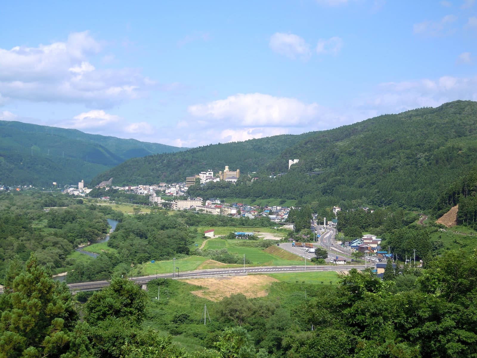 Naruko Onsen village panoramic view from hillside, green summer mountains, ryokan town below — photo by Yauchi, CC BY-SA 4.0