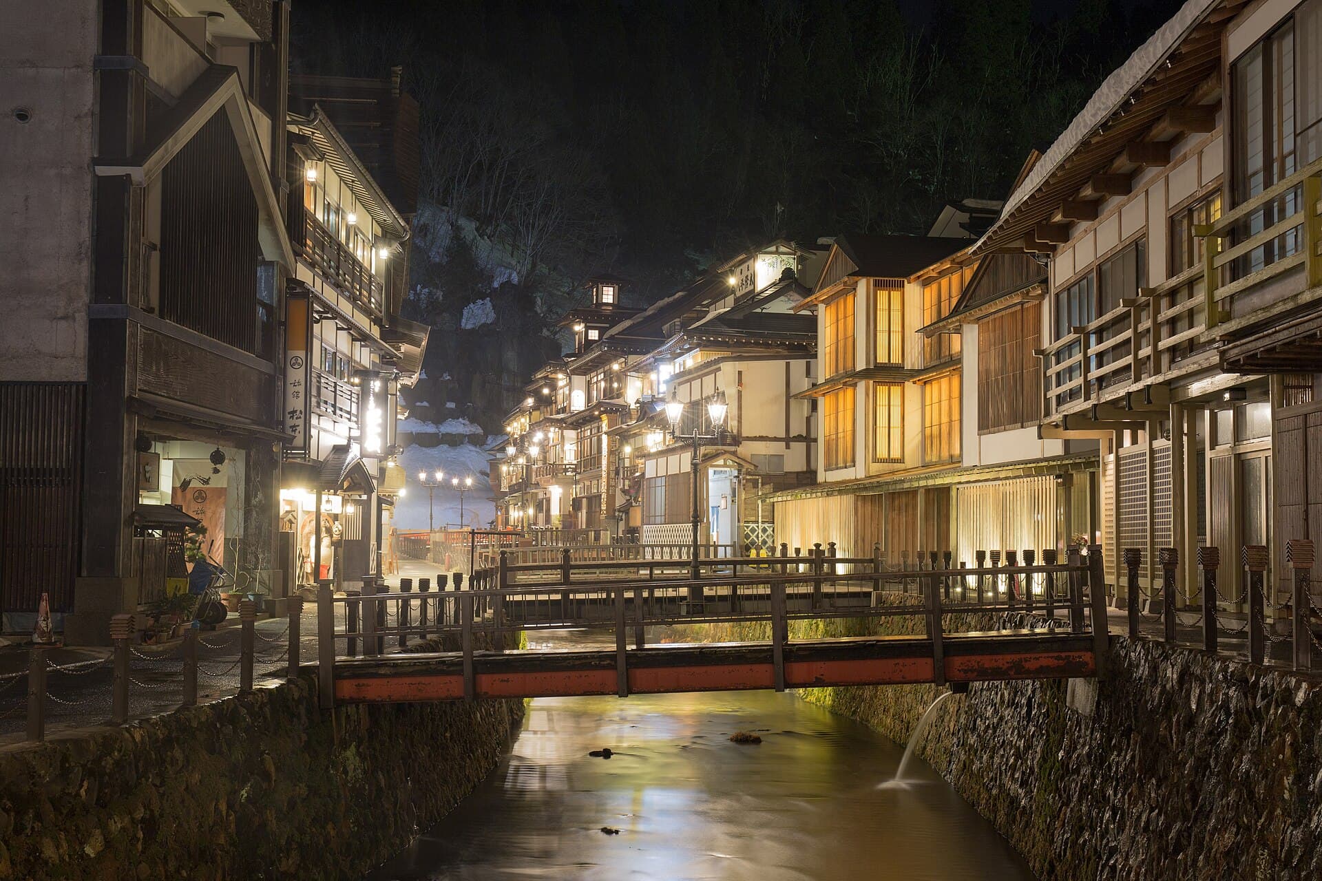 Snow-covered wooden ryokan buildings lining a narrow river gorge at a Japanese onsen town in winter, lit by warm light against deep snow — the visual atmosphere of Tohoku's hot spring towns