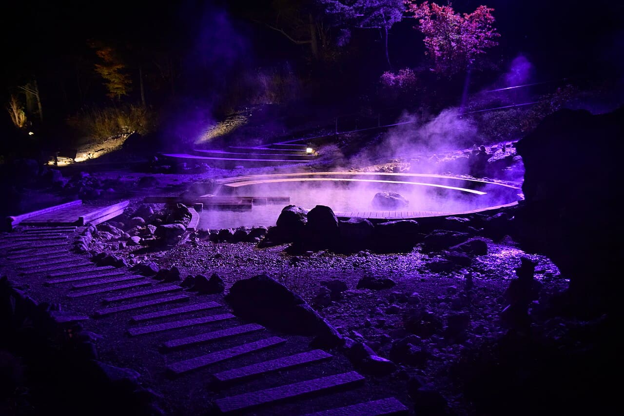 Steaming Yubatake hot-water field at the center of Kusatsu Onsen town at dusk, lined with wooden ryokan buildings — the iconic budget onsen anchor of central Japan