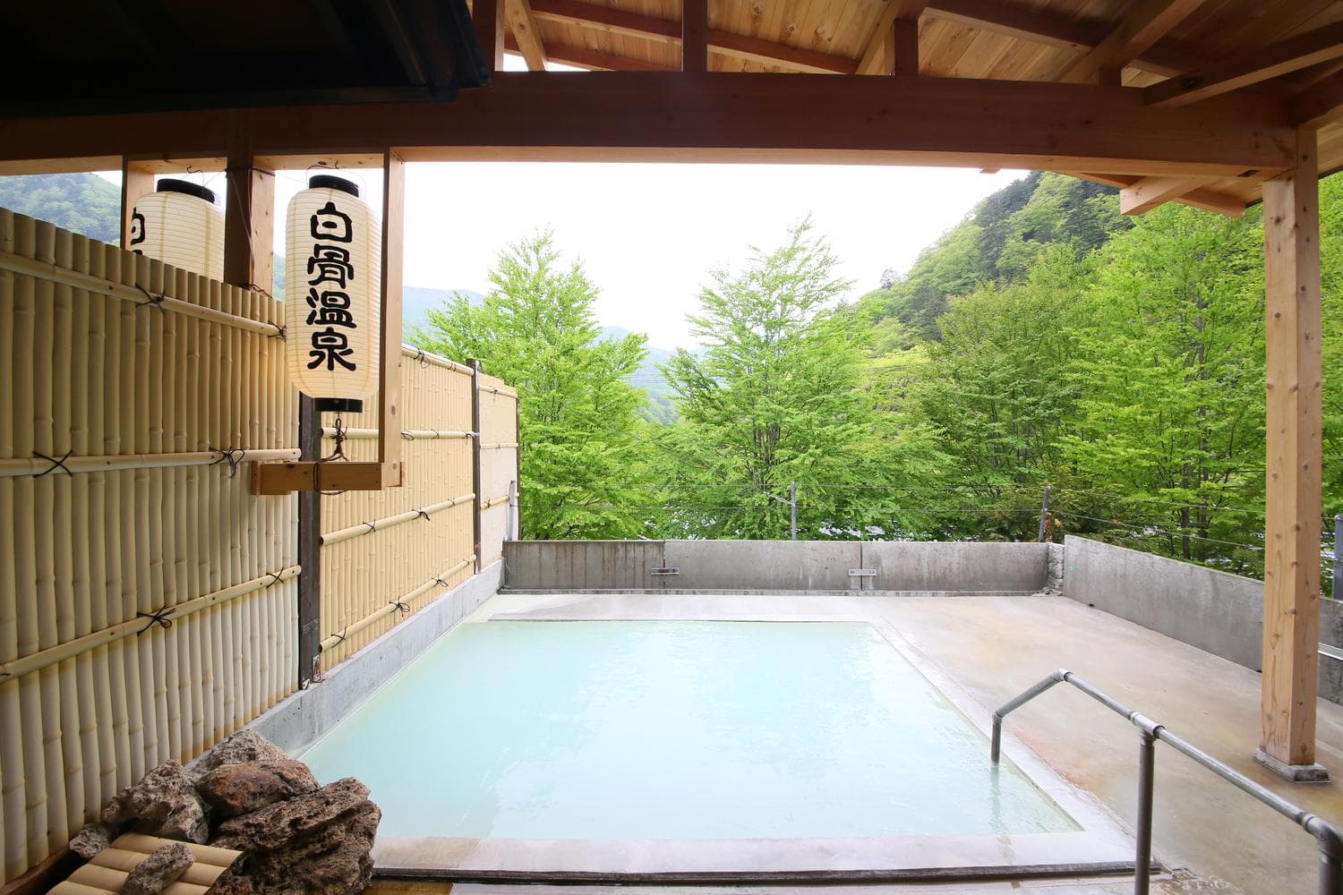 Open-air rotenburo bath with bamboo screen and lit lanterns overlooking green mountains at Shirahone Ebisuya