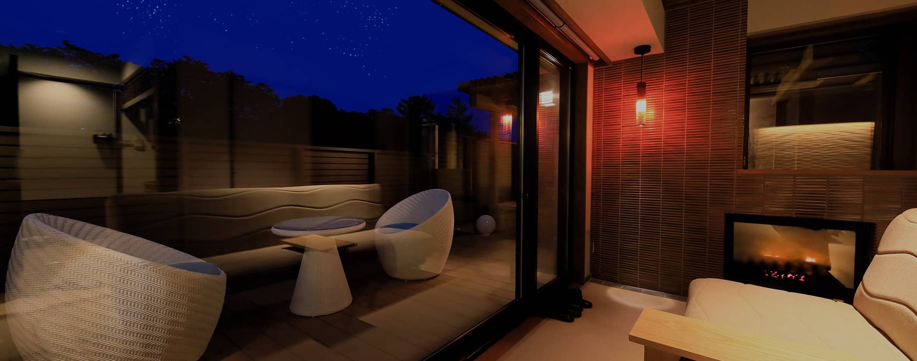 Guest room private terrace at dusk with rattan lounge chairs, fireplace and starry night sky beyond glass