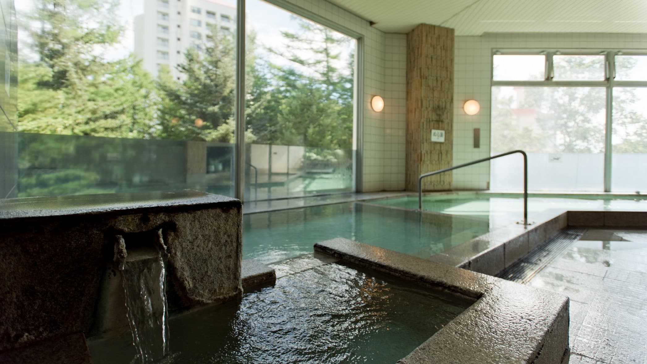 Indoor hot spring bath at Hotel Village with tiled pools and large windows looking onto green trees