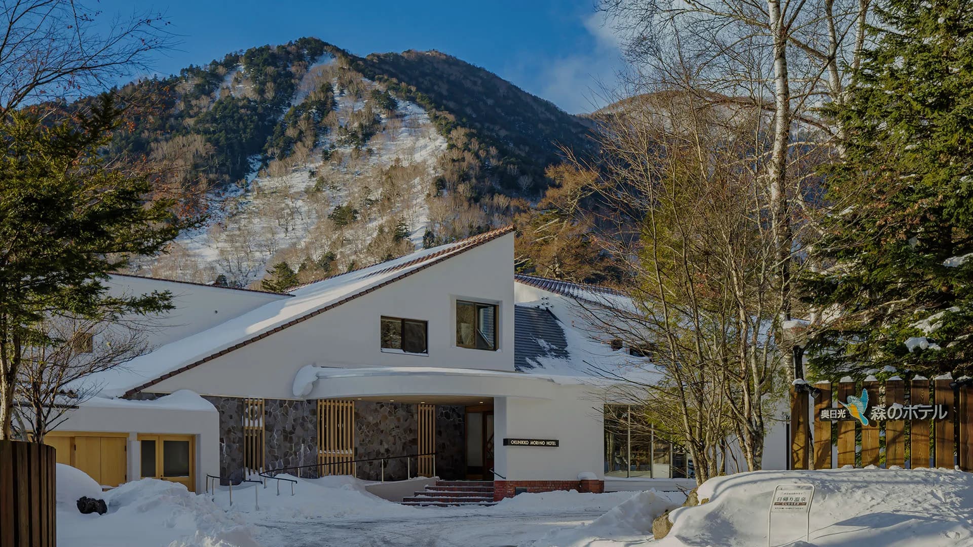 Snow-covered exterior of Oku Nikko Mori no Hotel beneath a forested Okunikko mountain on a clear winter day