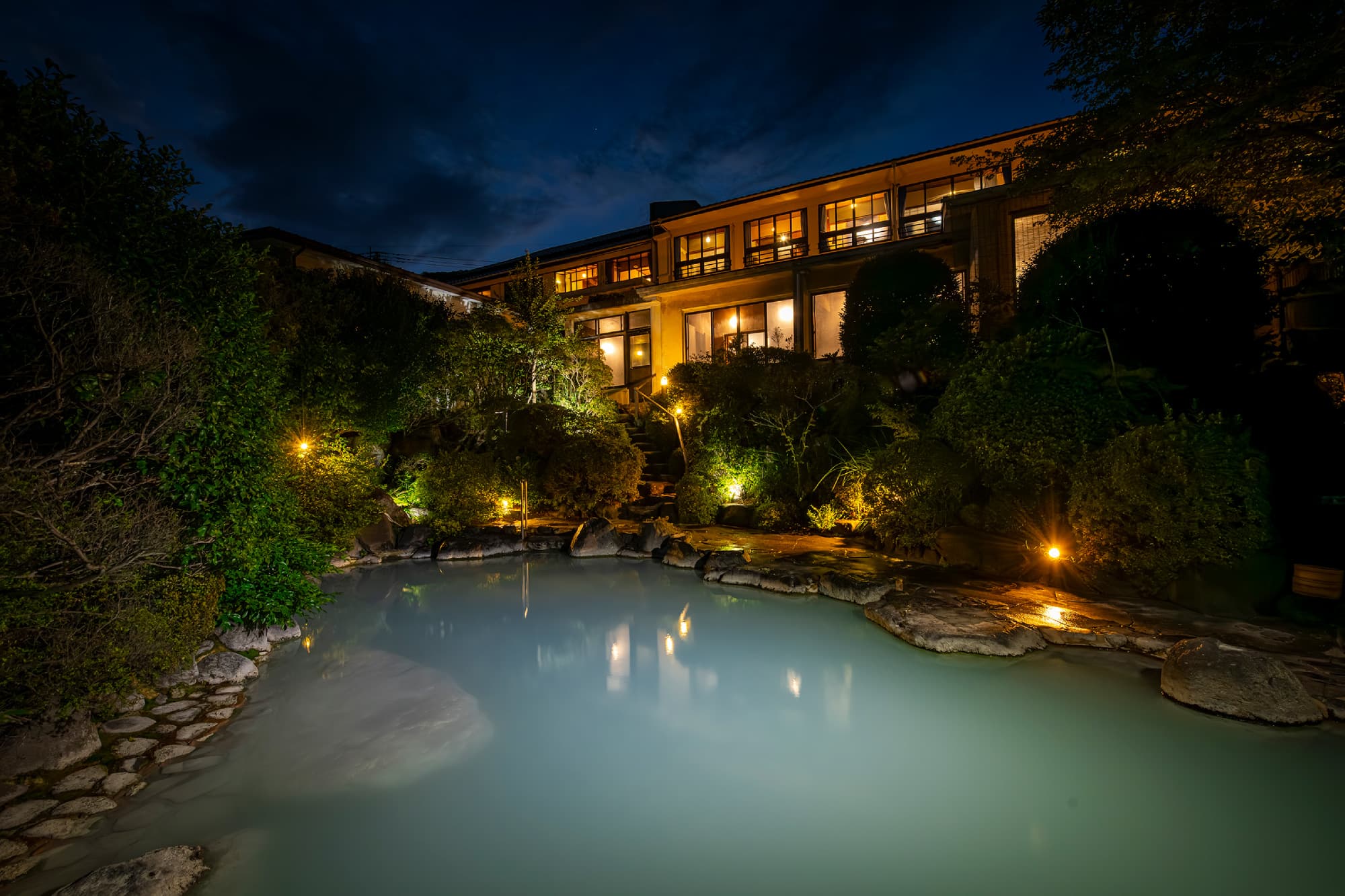 Pale blue garden rotenburo at night before the warmly illuminated main building and lantern-lit landscaped grounds, Okamotoya Ryokan, Myoban Onsen, Beppu
