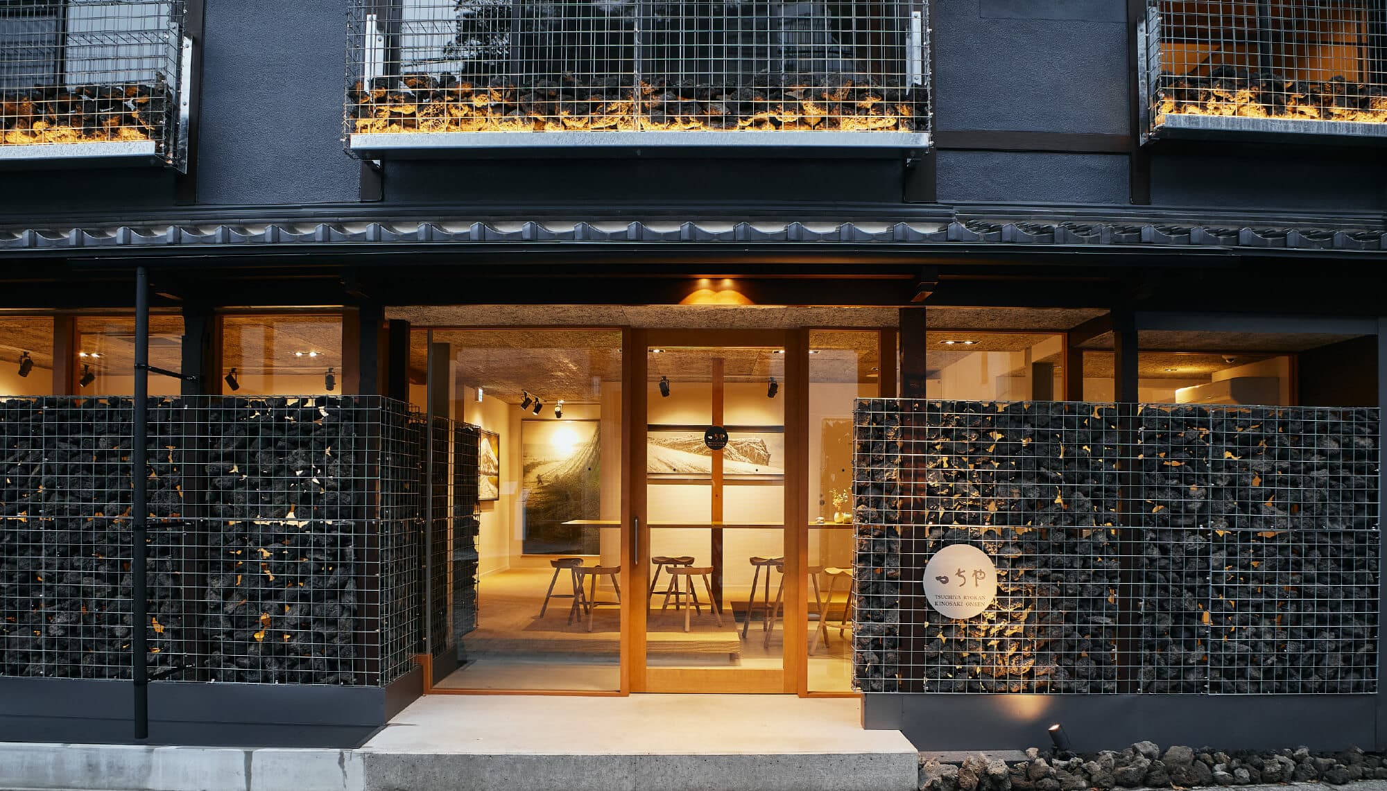 Tsuchiya ryokan entrance facade at dusk, dark timber exterior with warm interior lighting and stone gabion walls, Kinosaki Onsen