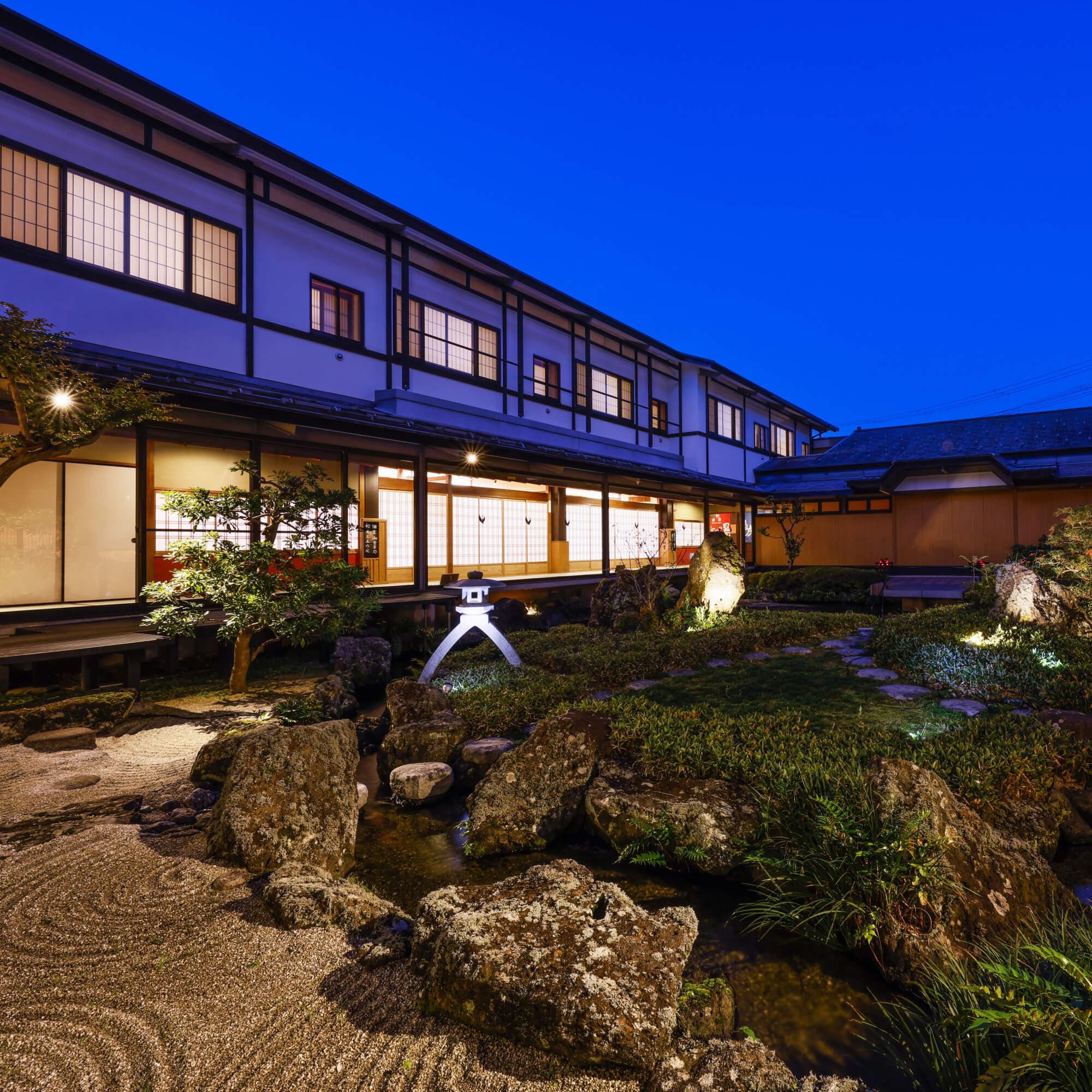 Traditional wooden ryokan facade overlooking a lantern-lit rock and gravel garden at twilight, Onishiya Suishoen, Kinosaki Onsen
