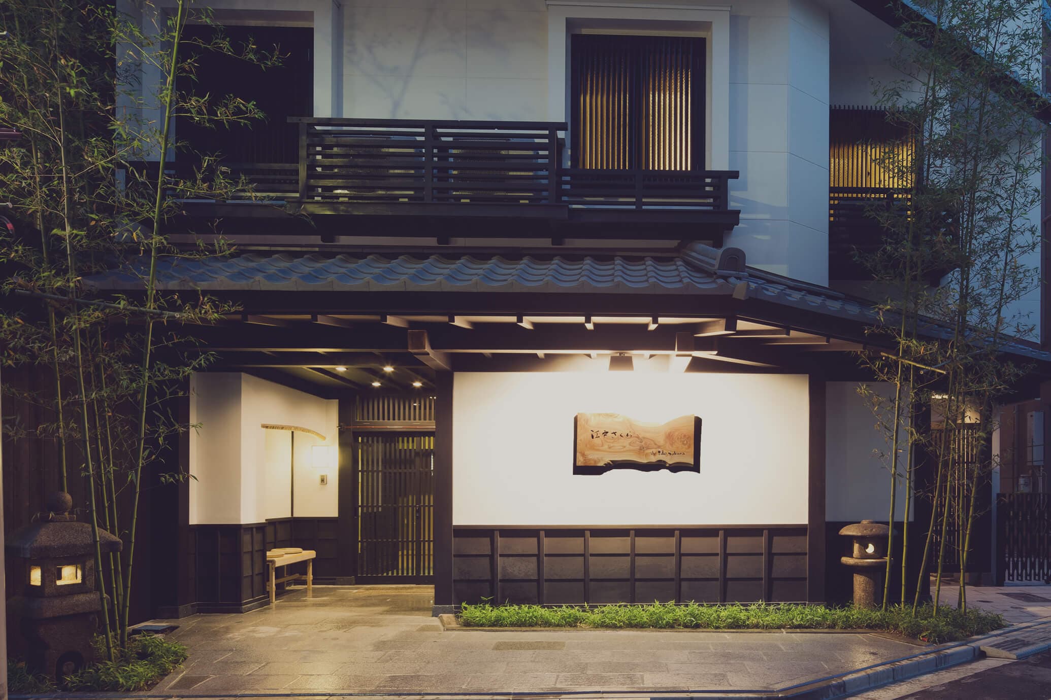 Illuminated wooden entrance and tiled-roof porch of The Edo Sakura inn at dusk framed by bamboo, Tokyo