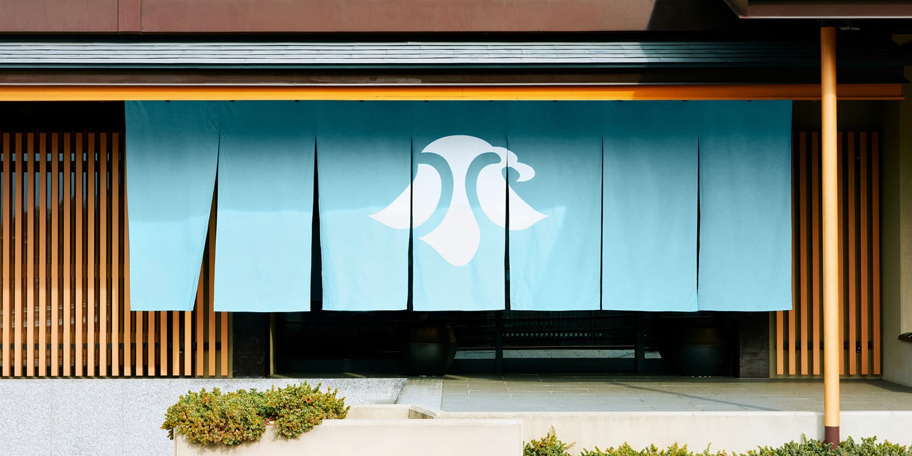 Entrance of Kozantei Ubuya with a pale-blue noren curtain bearing the inn's crest at the wood-slatted facade