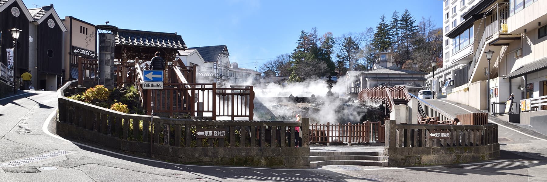 Panoramic daytime view of the Yubatake plaza streetscape with rising steam, wooden railings and the inn's neighbourhood