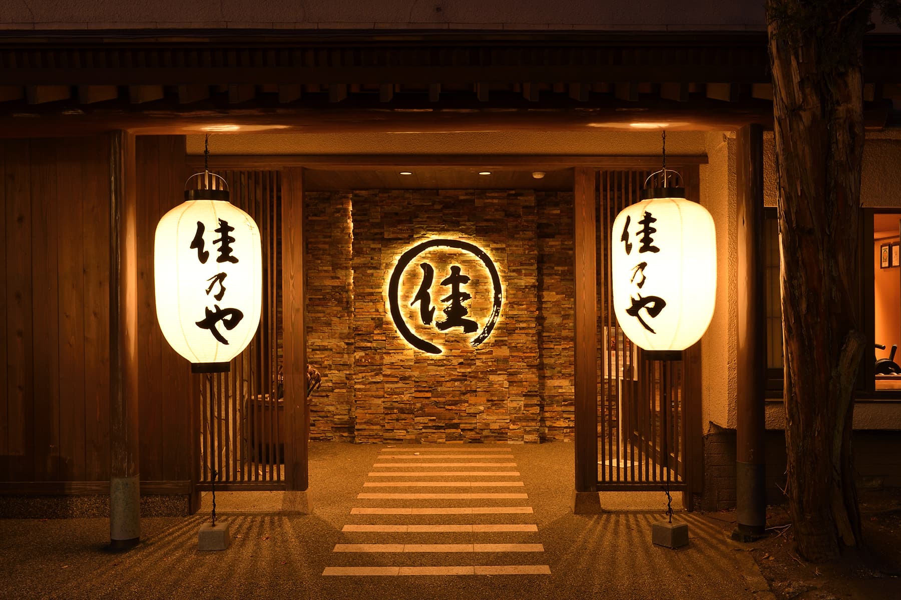 Illuminated wooden inn entrance at dusk flanked by two glowing paper lanterns over a stone-paved walkway