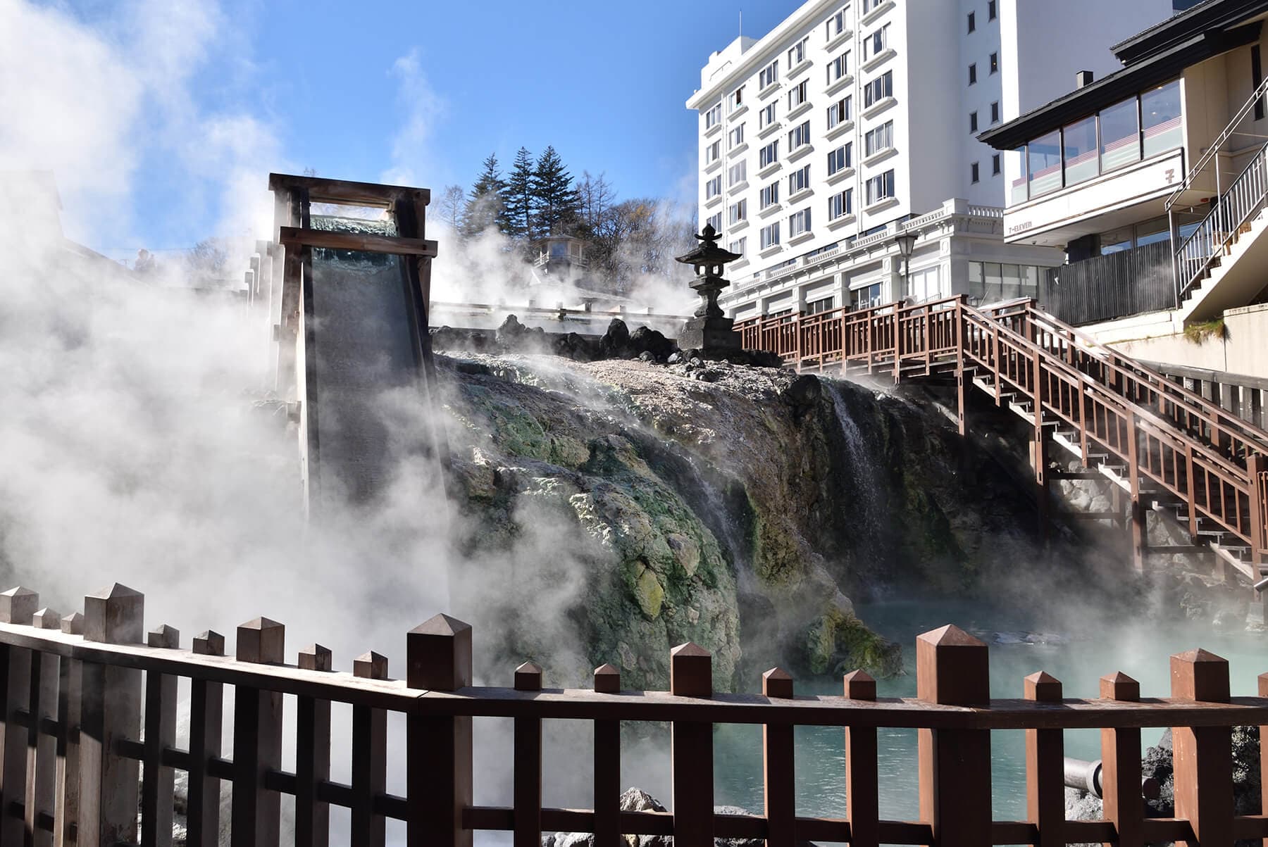 Steaming Yubatake hot-water field with wooden flumes and a stone lantern in central Kusatsu by day